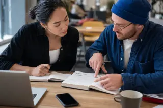 Focused freelancers sharing handwritten notes at a wooden table in a sunlit coworking space, engaged in thoughtful discussion.
