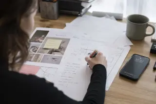 Freelancer reviewing handwritten client notes at a home desk, surrounded by documents and soft natural light.