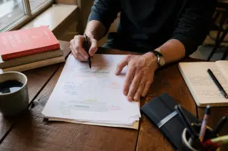 Freelancer's hands reviewing annotated printouts and notes on a wooden café table near a window with soft natural light.