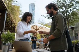 Two freelancers exchange business cards at an outdoor table in a bright, relaxed coworking terrace with a natural, urban backdrop.