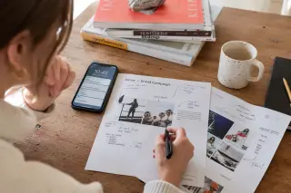 Freelance communication strategist reviewing marked-up documents at a wooden desk in a sunlit, lived-in home office, with smartphone and magazines nearby.